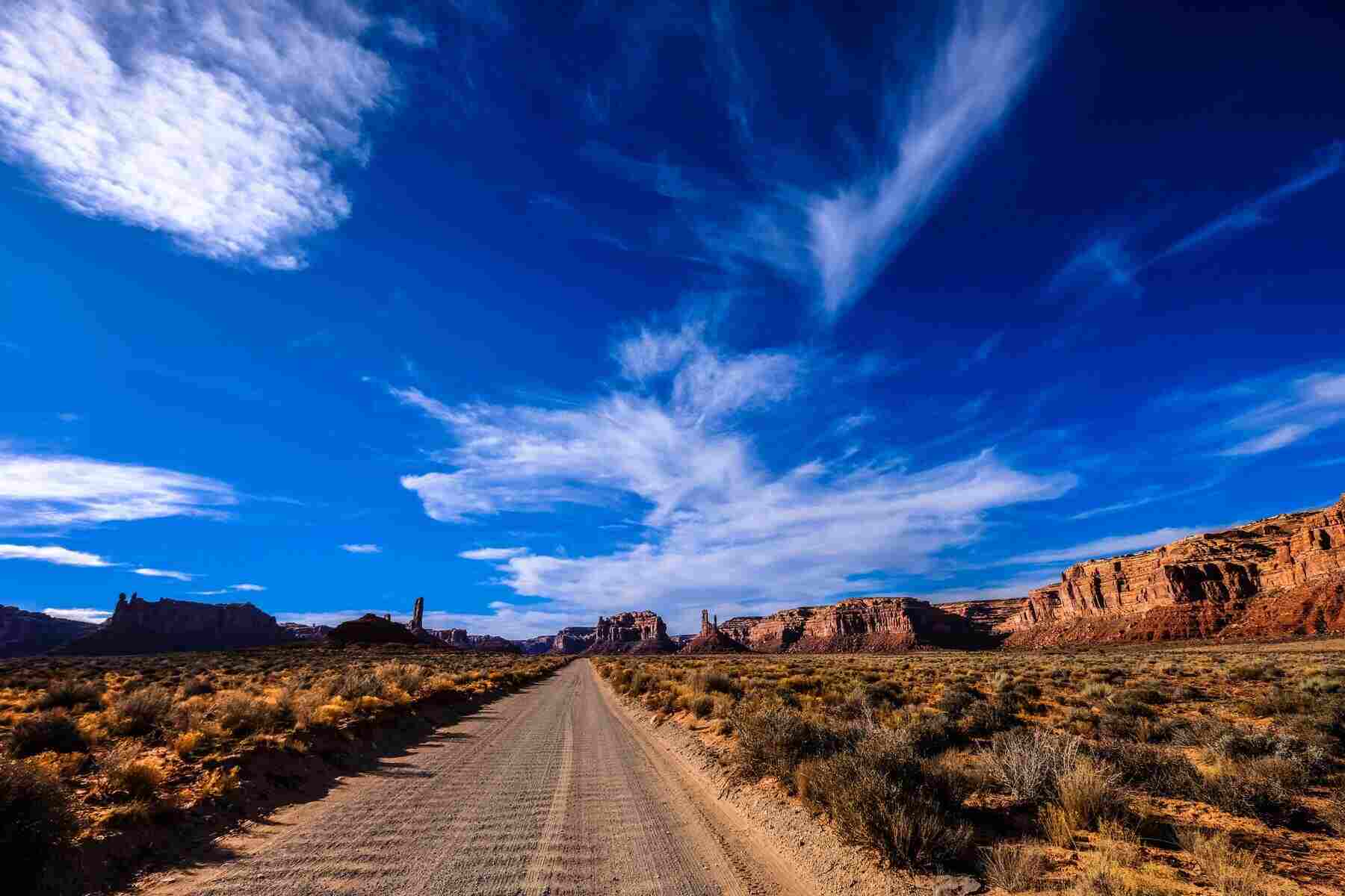 A long road in the desert. The sky is blue with clouds, and the surrounding dry landscape is orange.