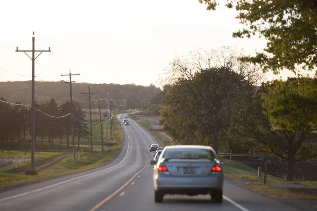 Rear view of cars driving along a winding rural road at sunset, with power lines and trees on both sides.